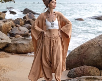 May include: A woman wearing a light brown, loose-fitting, long-sleeved top and matching trousers. She is standing on a rock by the ocean. The top has a wide, flowing silhouette and the trousers are gathered at the waist.