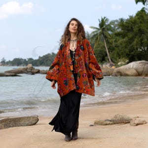 May include: A woman wearing a long-sleeved, rust-coloured velvet kimono with a floral print. The kimono has a wide, flowing silhouette and is worn over a black maxi skirt. The woman is standing on a sandy beach with the ocean in the background.