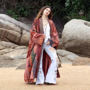 May include: A long, open-front patchwork kimono jacket featuring a variety of red, orange, and dark green fabric panels with intricate patterns. The garment is worn over a white sleeveless top and light-wash blue distressed denim flare jeans.
