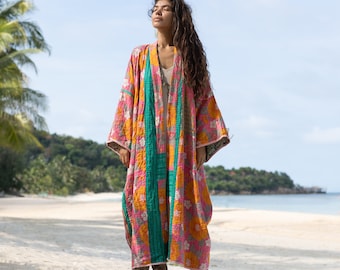 May include: A long, colorful kimono-style robe with a floral pattern in pink, orange, and gray. The robe has a teal panel down the front and is worn on a sandy beach. The sleeves are long and the robe reaches the ankles.