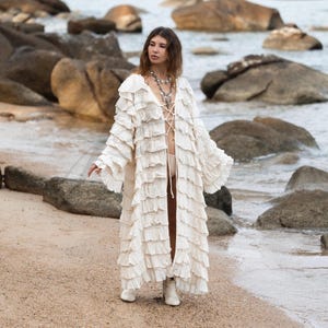 May include: A cream-colored, long, ruffled duster coat with a lace-up closure. The coat has wide sleeves and is worn with white boots. The background features a beach with rocks and the ocean.