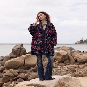 May include: A long-sleeved, open-front jacket featuring a dark black base with a geometric pattern in deep red and white. The garment has a V-neckline, button closures, and a loose, relaxed fit, paired with dark blue denim flare-leg jeans.