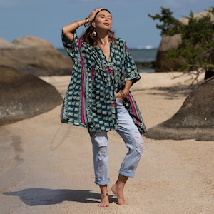May include: A teal, patterned tunic with a V-neck and wide sleeves. The tunic has a floral design with pink accents. The model is wearing light blue, ripped jeans. The background is a beach with rocks and the ocean.