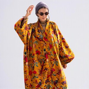 Close-up front view of the open-front velvet jacket. The rich golden mustard velvet has a soft sheen. Detailed view of the "Golden Sunbird" print featuring perching birds and blossoming flowers. Model wears layered boho jewelry and sunglasses, emphasizing the luxe, resort-wear style of the Mai collection.
