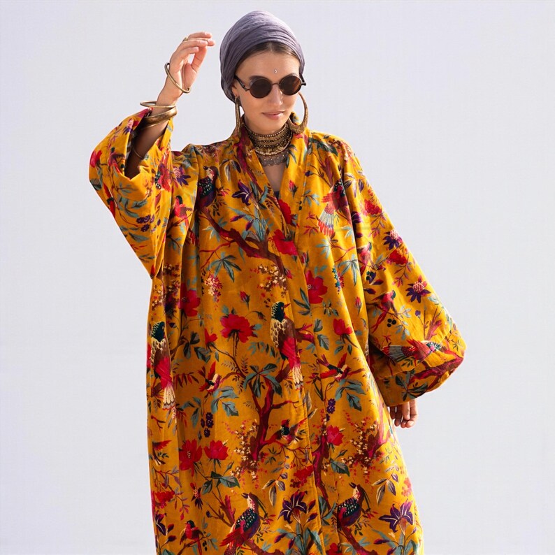 Close-up front view of the open-front velvet jacket. The rich golden mustard velvet has a soft sheen. Detailed view of the "Golden Sunbird" print featuring perching birds and blossoming flowers. Model wears layered boho jewelry and sunglasses, emphasizing the luxe, resort-wear style of the Mai collection.