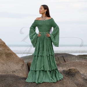 Lucia Boho Maxi Dress in Elderfern Green is a handmade cotton wedding gown featuring an off-shoulder neckline, smocked bodice, and layered ruffle skirt. Designed for beach brides, elopements, and goddess ceremonies, this dress blends vintage bell sleeves with breathable raw cotton for comfort and elegance. Perfect for plus-size women, maternity photoshoots, or barefoot rituals under the trees.