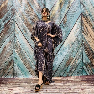 May include: A dark blue kaftan dress with a beige striped pattern. The dress has wide sleeves and a flowing silhouette. The model is wearing black platform sandals. The background is a wooden wall with a blue and brown color scheme.