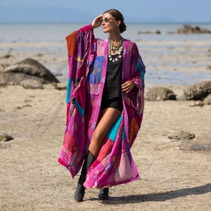 Puede incluir: Un largo kimono patchwork en tonos rosa, morado y azul. El kimono presenta una variedad de telas estampadas y se usa con una camiseta negra y botas. La persona lleva gafas de sol y collares.