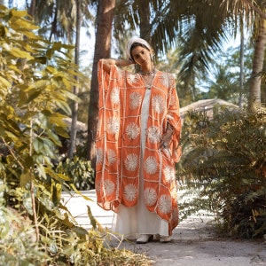 May include: A woman wearing a long, orange kimono with white embroidered flowers. The kimono has a loose fit and is worn over a white dress. The woman is standing in a tropical garden setting.