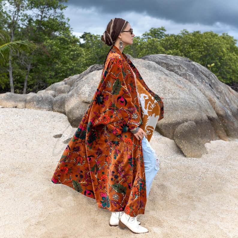 Side view of the Amber Flora Harper Jade Velvet Kimono Robe, photographed against a dramatic rocky coastal landscape with dark clouds above. The floor-length velvet duster sways in motion, revealing cream tribal-inspired lining details and a rich tapestry of jewel-toned florals on a burnt orange base. Designed for plus size inclusivity and fall boho elegance, this handcrafted robe combines artisan detail with modern styling, layered here over denim and boots.