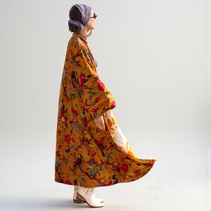 Side profile of the long velvet kimono coat in motion. The heavy, lined cotton velvet fabric flows gracefully, highlighting the ankle-length cut and wide kimono sleeves. The golden yellow background contrasts with the vibrant red floral and bird print. The image demonstrates the high-quality drape and substantial weight of the garment.