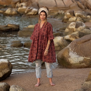 Puede incluir: Una mujer con un vestido granate de manga corta con estampado floral, combinado con vaqueros desgastados de color azul claro. También lleva una diadema color crema y pulseras doradas. El fondo muestra una playa con rocas y agua.