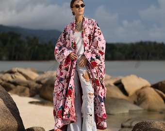 May include: A long, flowing pink floral kimono with red and blue accents. The kimono is worn over a white top and light blue ripped jeans. The background features a beach and water.