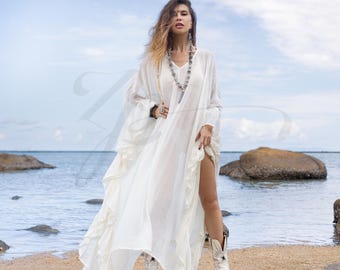 May include: A white, flowing maxi dress with ruffled detailing and long sleeves. The dress is worn by a person standing on a sandy beach with the ocean in the background.