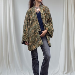 Model wearing an emerald green and gold hand block-printed velvet kimono jacket with wide sleeves, styled with jeans and brown boots, standing before soft white draped fabric background.