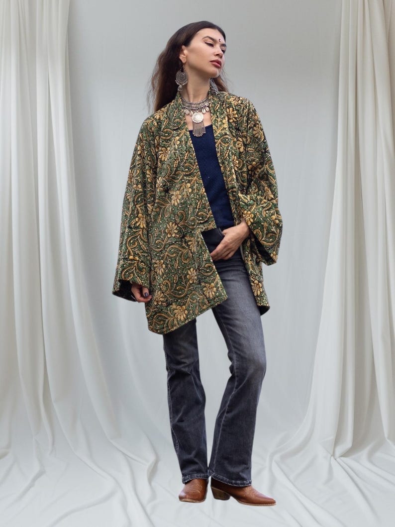 Model wearing an emerald green and gold hand block-printed velvet kimono jacket with wide sleeves, styled with jeans and brown boots, standing before soft white draped fabric background.