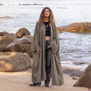 May include: A long, olive green open-front duster cardigan with wide sleeves, worn over a black sleeveless top and dark gray denim jeans. The garment features a loose, flowing silhouette that extends to the lower calf, made of a soft, matte fabric.