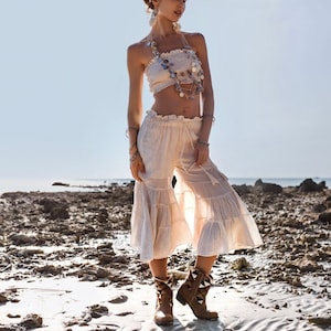May include: A woman wearing a white crop top with a beaded necklace and white pants with a ruffled hem. She is wearing brown boots with a pointed toe and a decorative design.