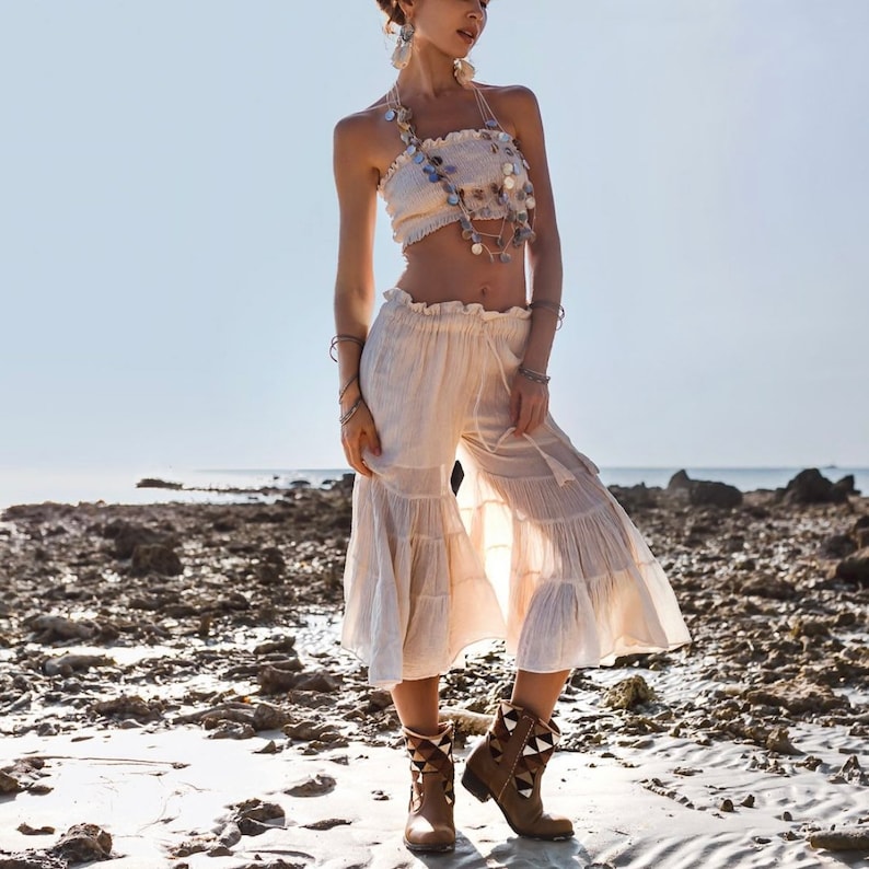 May include: A woman wearing a white crop top with a beaded necklace and white pants with a ruffled hem. She is wearing brown boots with a pointed toe and a decorative design.
