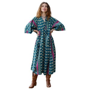2. Studio cutout image of the Cora Midi Dress in Peacock Rose against a white background. The oversized artisan cotton midi dress features a rich teal base with intricate floral-inspired motifs and vivid magenta side panels. Wide kimono sleeves, a flowing babydoll silhouette, and ankle-length drape create an effortlessly elegant bohemian shape styled with rustic brown boots.