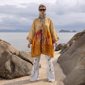 May include: A long-sleeved, loose-fitting tunic featuring a patchwork design in shades of yellow, gold, and red with a subtle repeating pattern. The garment is paired with light-wash, distressed wide-leg denim jeans with frayed hems.