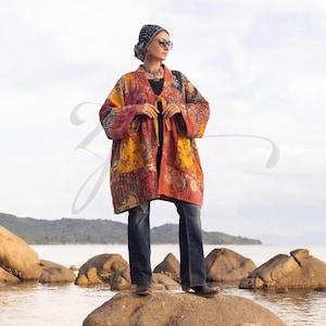 May include: A patchwork kimono-style jacket in shades of red, yellow, and orange. The jacket features a tie closure and long sleeves. The model is wearing the jacket with dark blue jeans and a patterned headscarf.