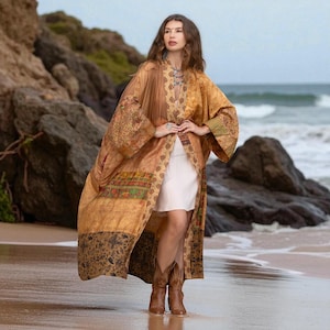 May include: A long, flowing, patterned kimono in shades of gold, brown, and green. The kimono features intricate floral and geometric designs. The garment is worn over a white dress and paired with brown cowboy boots. The background shows a beach and ocean.