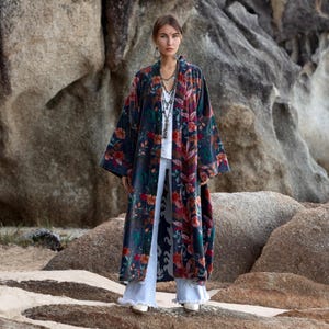May include: A long, flowing floral kimono in shades of navy, red, and green. The kimono features a vibrant floral pattern and is worn over a white top and light blue flared jeans. The outfit is completed with white shoes.