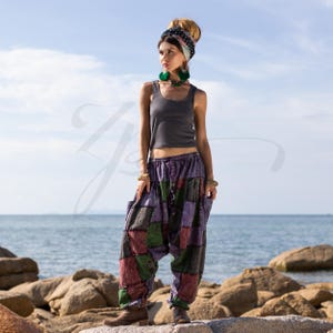 May include: A woman wearing a gray tank top and patchwork pants with a colorful pattern. She is standing on a rocky beach with the ocean in the background.