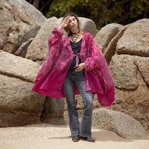 Puede incluir: Una chaqueta estilo kimono de color magenta vibrante con un patrón floral dorado detallado, mangas largas y holgadas, y un cierre de lazo frontal. La prenda se lleva sobre una camiseta negra sin mangas y vaqueros gris oscuro, con botines de cuero.
