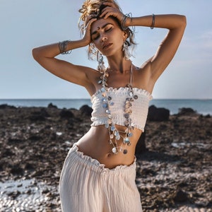 May include: A woman wearing a white strapless top with a white skirt and a silver necklace with coin-shaped charms. She is wearing several silver bracelets on her wrists and has her hair pulled back.