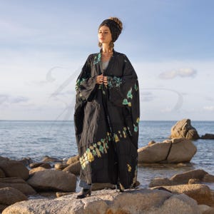 May include: A long black tie-dye kimono with green and yellow accents. The kimono is worn by a person standing on a rocky beach.
