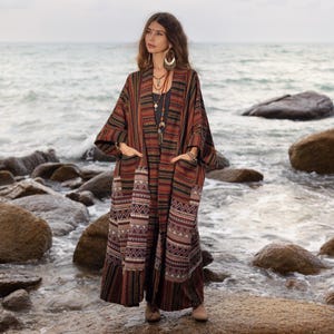 May include: A long, flowing, red, black, and white patterned kimono with long sleeves and pockets. The kimono is worn by a person standing on a rocky beach.