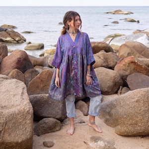 May include: A loose-fitting, button-down tunic in a deep purple hue with a subtle, shimmering floral pattern. The garment features three-quarter length sleeves, a V-neckline, and a gathered waistline that creates a flared, flowing silhouette.