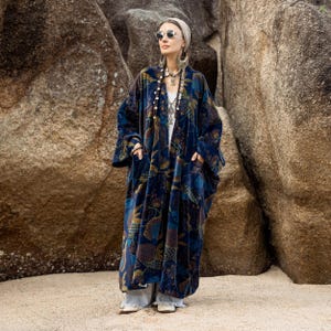 Könnte beinhalten: Langer, fließender marineblauer Samtrobe mit einem Blumenmuster in Gold- und Türkistönen. Der Robe hat weite Ärmel und Taschen. Die Person trägt eine Sonnenbrille und Schmuck.