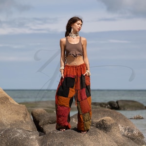 May include: Patchwork pants in shades of red, orange, black, and green. The loose-fitting pants have a drawstring waist and pockets. The model is wearing a brown crop top and standing on a rock near the ocean.