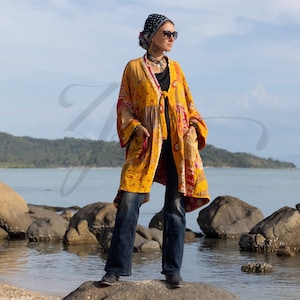 May include: A long, yellow patchwork kimono-style jacket with red and floral accents. The jacket is worn over a black top and dark blue jeans. The person is standing on a rock near the water, wearing sunglasses and a patterned headscarf.