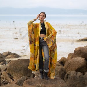 Puede incluir: Un kimono largo abierto por delante con un diseño de patchwork en varios tonos de amarillo, dorado y mostaza con patrones intrincados. La prenda tiene mangas anchas y fluidas y un dobladillo recto, usado sobre un top corto negro y jeans grises.