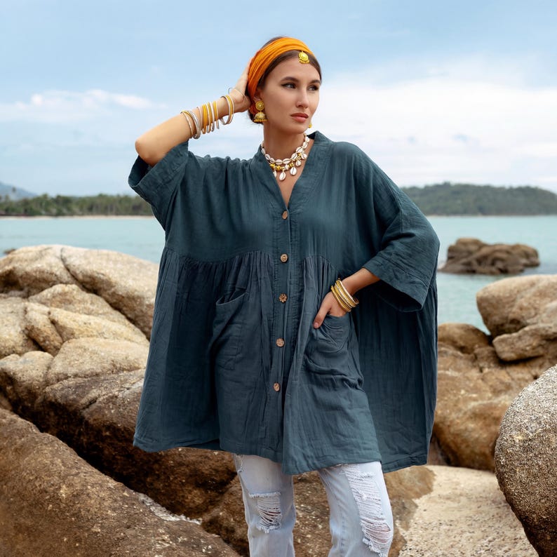 Three-quarter close-up of the Aria Tunic in Smoky Jade cotton gauze highlighting the wooden button front, deep functional pockets, and textured crinkle cotton fabric. The oversized boho silhouette and wide sleeves create an airy drape. Styled with layered jewelry and rolled denim jeans against a coastal backdrop, the tunic embodies relaxed luxury and effortless bohemian elegance.