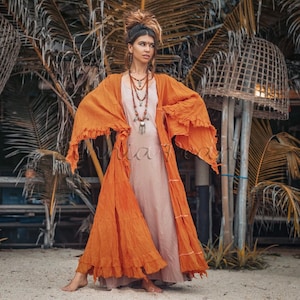 May include: A woman wearing a long, flowing orange dress with a ruffled hem. The dress has a light brown under layer and is worn with a long, layered necklace with silver and red beads. The woman is standing in front of a wooden structure with palm trees in the background.