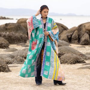 Puede incluir: Una chaqueta tipo kimono larga y abierta por delante, confeccionada con tela de retazos multicolor con intrincadas costuras kantha. La prenda muestra una mezcla vibrante de patrones florales y geométricos en azul, amarillo, rosa y turquesa.
