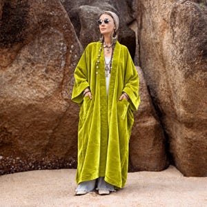 May include: A long, chartreuse velvet duster coat featuring an open front, wide sleeves, and two large patch pockets. The garment is worn over a white top and light-colored wide-leg trousers, accessorized with a head wrap, sunglasses, and jewelry.