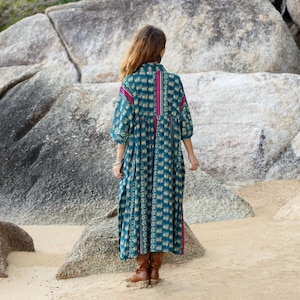 5. Back view of the Cora Midi Dress in Peacock Rose showing the full flowing silhouette and artisan print placement. The oversized midi dress features soft teal patterned cotton with contrasting magenta vertical accents and relaxed kimono sleeves. Styled outdoors among natural stone formations, the bohemian silhouette drapes elegantly with comfortable movement and effortless free-spirited style.