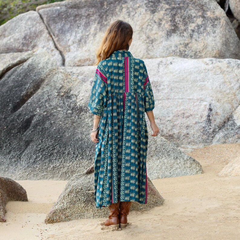 5. Back view of the Cora Midi Dress in Peacock Rose showing the full flowing silhouette and artisan print placement. The oversized midi dress features soft teal patterned cotton with contrasting magenta vertical accents and relaxed kimono sleeves. Styled outdoors among natural stone formations, the bohemian silhouette drapes elegantly with comfortable movement and effortless free-spirited style.