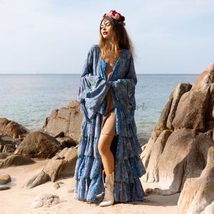 May include: A blue floral maxi dress with a ruffled skirt and long sleeves. The dress is worn by a person with a flower crown on their head. The person is standing on a beach with rocks in the background.