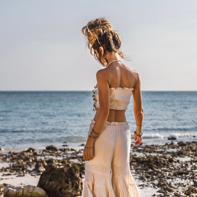 May include: A woman wearing a white strapless top with a shell embellishment and white wide-leg pants with a ruffled hem. She is standing on a rocky beach with the ocean in the background.