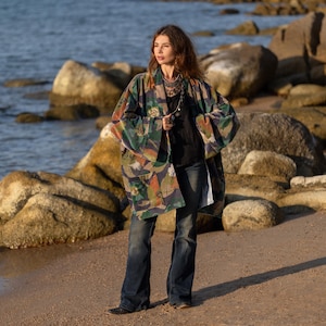 May include: A patterned kimono in shades of green, brown, and orange. The kimono is worn over a black top and blue jeans. The background features a beach with rocks and water.