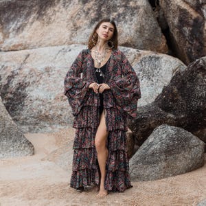 May include: A long, tiered, patterned kimono in shades of black, red, and teal. The kimono has wide sleeves and a front slit, worn over a black swimsuit. The model is standing on a sandy beach with large rocks in the background. Bohemian style.