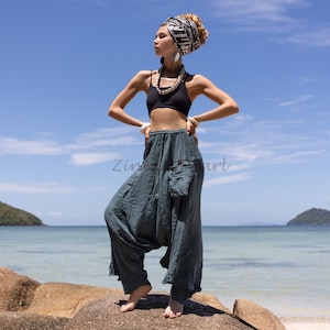 May include: A woman wearing a black crop top, a patterned headscarf, and loose-fitting, teal-colored pants with a large pocket on the left side. She is standing on a rock with the ocean and sky in the background.