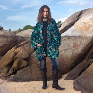 May include: A teal velvet kimono-style jacket with a floral pattern in shades of orange and green. The jacket is worn over a black top and paired with dark wash jeans and black knee-high boots.